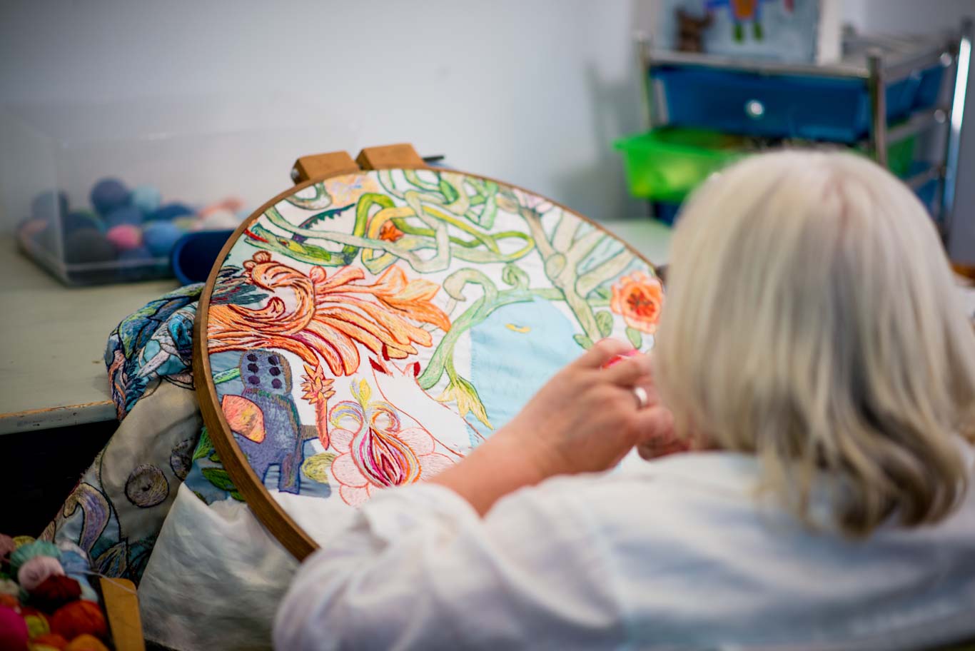 Anna Torma, facing away from the camera is seated at her worktable, a large embroidery hoop is sitting on the table in front of her. In the embroidery hoop is a work in progress full of multiple different colors of embroidery thread.