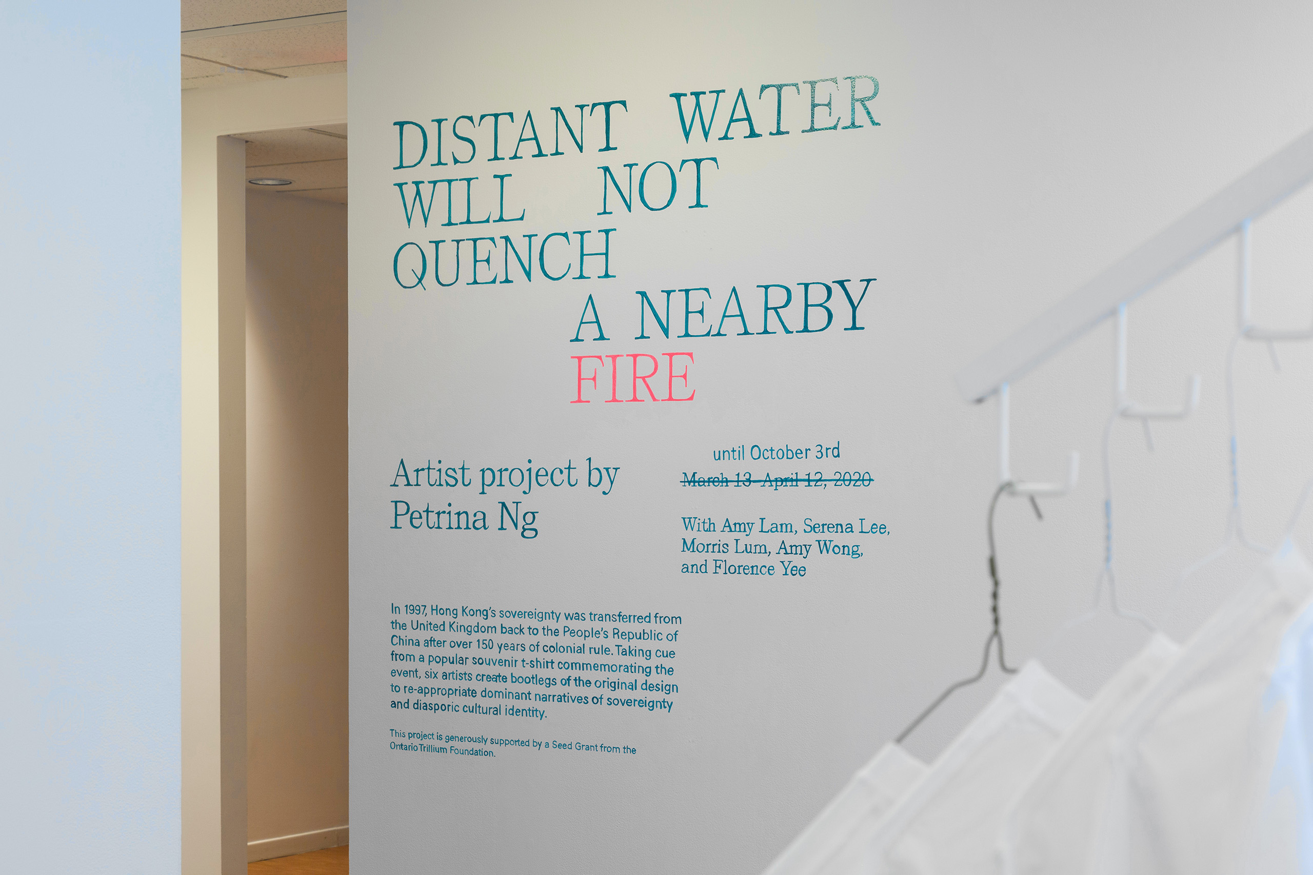 Exhibition intro text painted in teal and pink on a white wall. The edge of hanging t-shirts is visible in the bottom right