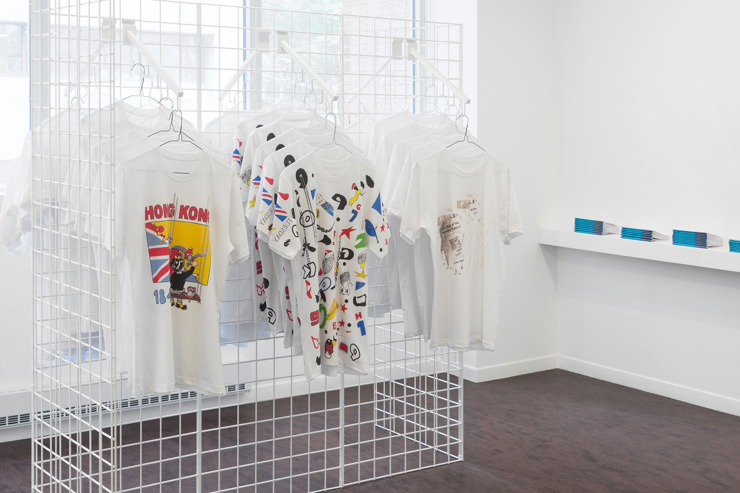 T-shirts hanging on a white wire frame, with stacks of booklets on a narrow ledge behind them