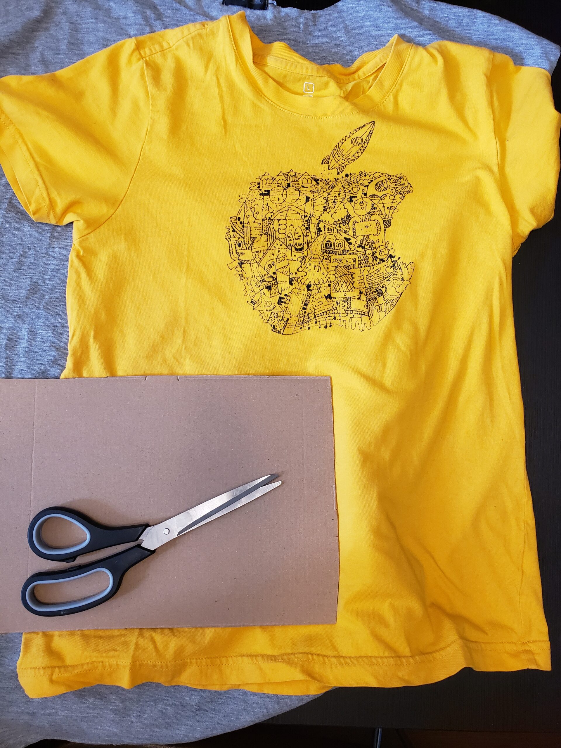 A yellow t-shirt, a rectangular piece of cardboard and a pair of scissors are laid on a table