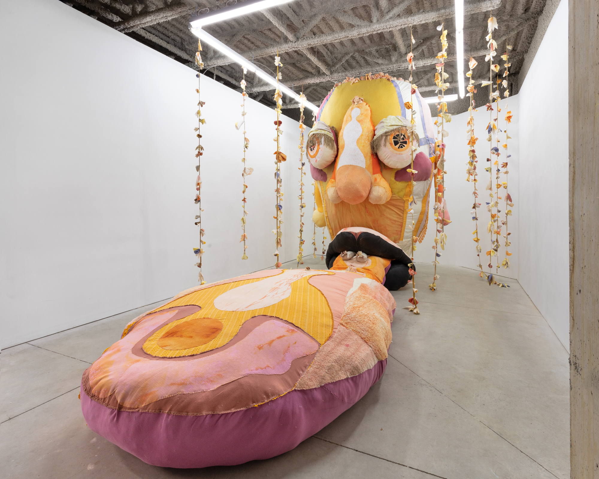 Image shows a large textile sculpture in a white cube space. The structure is a larger than life, expressive human head built from textiles. The head has a disproportionately large tongue extending out from its lips. The tongue stretches out across the floor. The head and tongue are colourful; pinks, yellows, blacks, whites and purples. Hanging around the head and tongue are long flower ropes suspended from the ceiling.