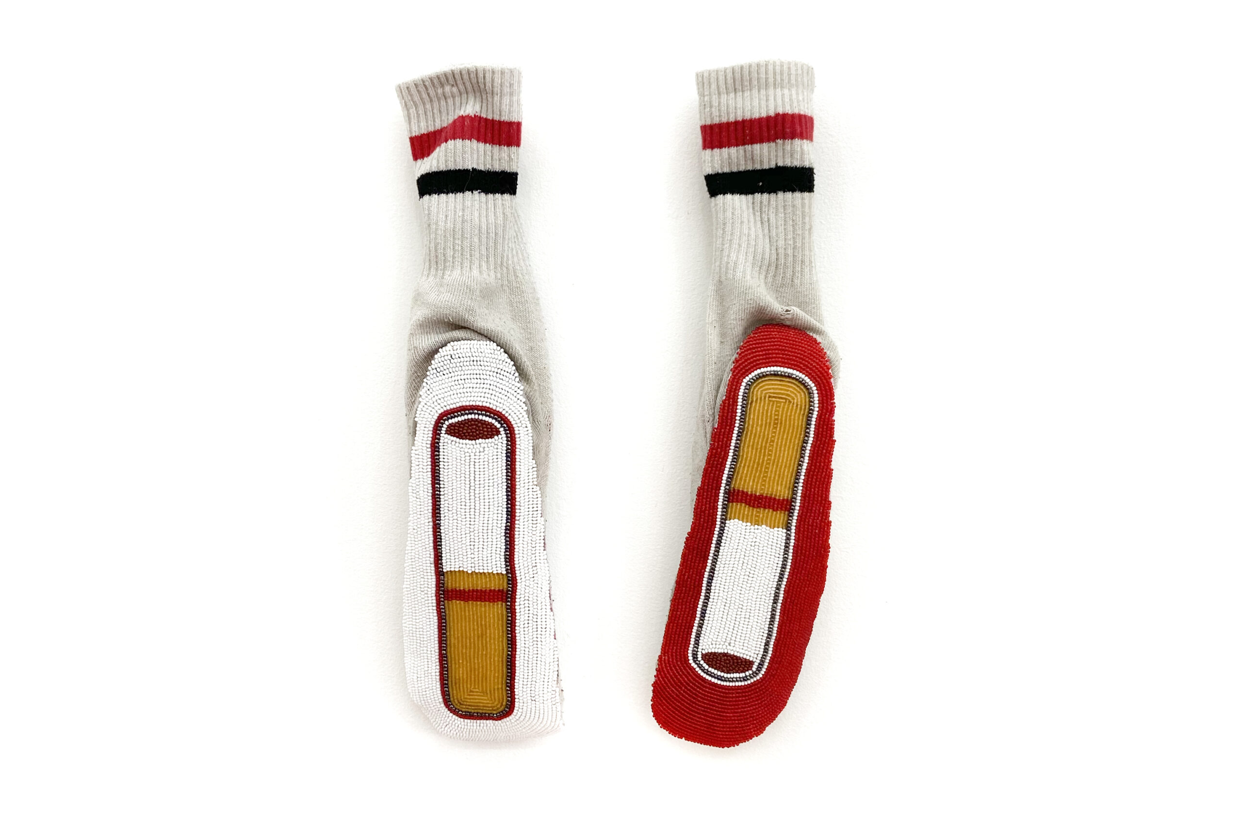An image of Audie Murray's piece Tabacco Offering. The image shows a pair of white socks. The bands of the sock have two stripes: one red, one black. The soles of the socks face the camera. The sock soles are richly beaded in red, ochre, white and black.