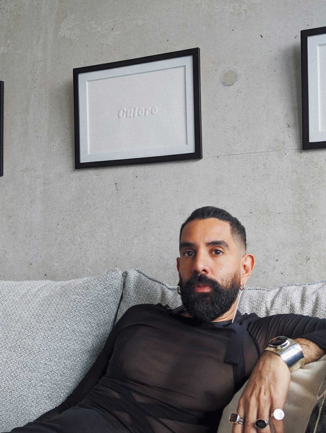Armando Perla sitting on a white couch against a white wall. Armando has a dark coloured beard and short dark hair. They are wearing a black sheer top with solid black crisscrossing ties. They are wearing a silver watch and rings while leaning against the arm of the couch.